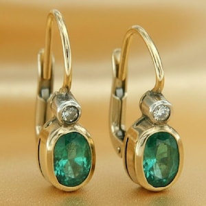 Puede incluir: Un par de pendientes dorados con piedras preciosas ovaladas de color verde esmeralda. Cada pendiente presenta una pequeña piedra redonda y transparente encima de la gema verde. Cierre de palanca, sobre un fondo dorado.