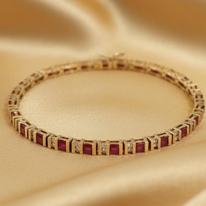 May include: A gold bracelet with alternating rectangular red gemstones and small white gemstones. The bracelet is set against a gold satin background, creating a luxurious aesthetic. The clasp is visible at the top of the bracelet.