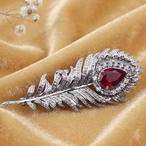 Puede incluir: Un broche de pluma en tono plateado con una prominente piedra preciosa de color rojo intenso en forma de pera en el centro, rodeada por un halo de pequeñas piedras transparentes engastadas en pavé. La superficie está incrustada con cristales.