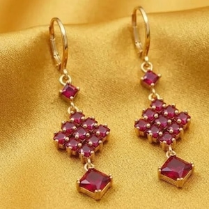 May include: A pair of gold-tone drop earrings featuring a lever-back closure. Each earring displays a small square red gemstone at the top, followed by a cluster of nine square red gemstones arranged in a grid, and a single square red gemstone drop.