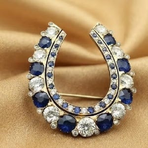 Peut inclure: Une broche en forme de fer à cheval ornée de pierres rondes bleu foncé et de pierres blanches transparentes alternées. Le bord extérieur est bordé de pierres plus grandes, tandis qu'une rangée intérieure de petites pierres bleues suit la courbe.
