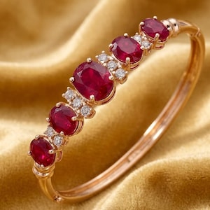 May include: A gold bangle bracelet featuring oval-cut rubies and small diamonds. The rubies are a deep red color and are set in a row along the top of the bracelet. The diamonds are clustered between the rubies.