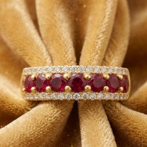 Op de afbeelding: Een gouden ring met een centrale rij van zeven rondgeslepen dieprode robijnen, geflankeerd door twee buitenste rijen van kleine, heldere, rondgeslepen diamanten. De ring is gezet in een gepolijste geelgouden band met een zetting voor meerdere stenen.