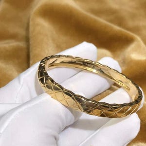 May include: A polished gold bangle bracelet featuring a repeating quilted diamond pattern across its exterior surface. The interior of the band displays engraved text, including the brand name Chanel, visible within the smooth metal finish.