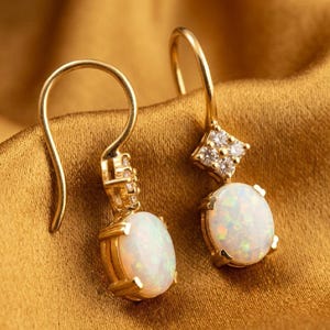 Pode incluir: Um par de brincos pendurados dourados com pedras de opala ovais. Os brincos têm um detalhe em forma de diamante com pequenas pedras transparentes. As opalas têm uma base branca leitosa com flashes de cor iridescente.