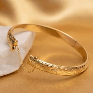 Puede incluir: Una pulsera rígida articulada de tono dorado con un patrón de volutas grabado en la superficie exterior. La pulsera incluye una delicada cadena de seguridad que conecta los dos extremos, asegurados mediante un mecanismo de bisagra funcional.