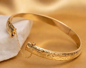 May include: A gold hinged bangle bracelet featuring an intricate engraved scrollwork pattern along the outer surface. The bracelet includes a delicate gold safety chain connecting the two ends, which are secured by a functional clasp mechanism.
