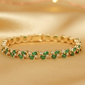 Puede incluir: Una pulsera dorada con diseño de hojas, con piedras preciosas de color verde esmeralda y transparentes. La pulsera está sobre un fondo dorado, resaltando el brillo y los intrincados detalles de la joya. El cierre es visible.
