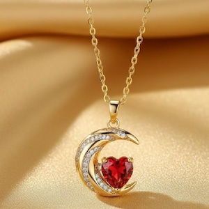 May include: A gold-colored necklace with a pendant featuring a red heart-shaped gemstone cradled within a crescent moon design. The crescent is embellished with small, clear gemstones. The chain and pendant are set against a gold-colored background.