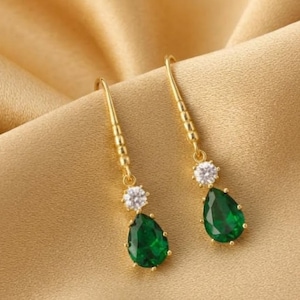 May include: A pair of gold drop earrings featuring a clear round faceted stone at the top and a larger pear-shaped emerald green faceted stone suspended below. The earrings have a thin gold hook wire and a polished gold metal setting.
