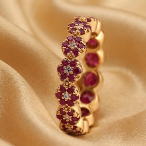 May include: A gold band ring featuring a continuous row of flower-shaped motifs. Each flower is crafted from six small, deep pink circular gemstones surrounding a single, tiny, clear white gemstone at the center of every floral cluster.