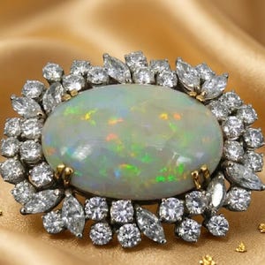 May include: An oval-shaped cabochon opal brooch featuring a central iridescent stone with flashes of green, orange, and yellow. The opal is surrounded by a halo of numerous round and marquise-cut clear stones set in a metallic frame.