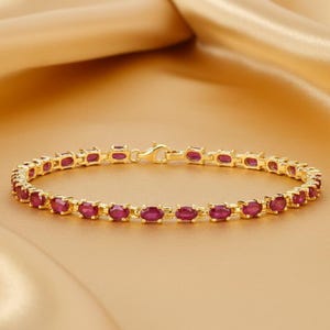 May include: A gold-colored bracelet featuring a series of oval-shaped, deep red gemstones. The bracelet is secured with a clasp. The gemstones are evenly spaced along the length of the bracelet, creating a continuous pattern. The jewelry is displayed on a gold-colored surface.