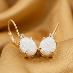 May include: A pair of gold-colored leverback earrings. Each earring features an oval, white opal stone with iridescent flecks, set in a gold frame. A small, clear gemstone is set above the opal. The earrings are displayed on a gold-colored fabric.