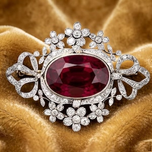 May include: An oval-shaped brooch featuring a large, deep red gemstone at the center. The piece is surrounded by a silver-toned metal frame encrusted with numerous small, clear, sparkling stones arranged in intricate floral and ribbon-like patterns.