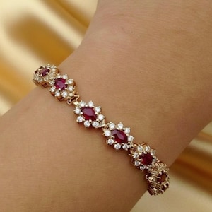 Könnte beinhalten: Goldfarbenes Armband mit ovalen roten Edelsteinen und kleinen, klaren Edelsteinen. Das Design des Armbands umfasst blütenartige Cluster, die ihm ein zartes Aussehen verleihen. Das Armband wird am Handgelenk getragen.