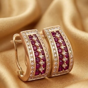 May include: A pair of gold hoop earrings featuring a central row of round, deep red rubies flanked by two rows of small, clear white diamonds. The earrings have a curved, rectangular shape with a hinged closure mechanism for secure attachment.