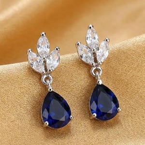 May include: A pair of silver-toned drop earrings. Each earring features a cluster of three clear, marquise-cut stones above a large, pear-shaped, deep blue gemstone. The earrings are set against a gold-colored fabric.