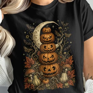 May include: A black t-shirt featuring a graphic print of a vertical stack of four carved jack-o'-lanterns. The design includes a crescent moon, moths, autumn leaves, and mushrooms in shades of orange, brown, and cream against the dark fabric.