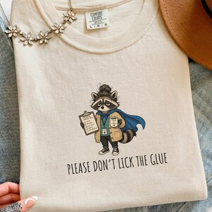 May include: An ivory t-shirt featuring a graphic of a raccoon wearing a blue cape, a tan cardigan, and a messy bun, holding a clipboard and a mug. The text below the raccoon reads, "PLEASE DON'T LICK THE GLUE." The shirt has a crew neckline.