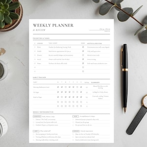 Puede incluir: Un planificador semanal minimalista con el texto "WEEKLY PLANNER & REVIEW" en negro. Un bolígrafo negro y un reloj de pulsera con una correa de cuero negro están a la derecha. Una pequeña planta suculenta y un tarro de cristal están a la izquierda.