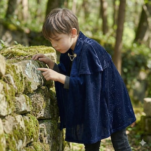 Op de afbeelding: Een donkerblauwe fluwelen cape met een gouden ketting sluiting. De cape wordt gedragen door een persoon in een bosrijke omgeving. De persoon wijst naar een met mos bedekte stenen muur.