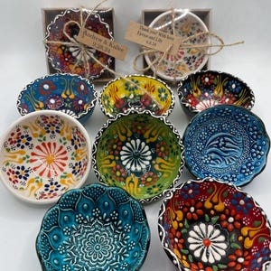 May include: A selection of vibrantly decorated ceramic bowls with floral designs. The bowls display a range of colours, including blue, red, yellow, and green, with white and black detailing. Some bowls are packaged in clear boxes with tags attached.