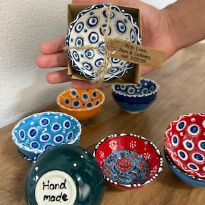 May include: A collection of small, hand-painted ceramic bowls in various colors and patterns. One bowl is in a cardboard box tied with twine and a tag that reads "With Love, Alan & Colleen est. 3/22/25". Other bowls are blue, orange, red, and green.