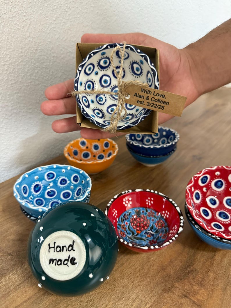 May include: A collection of small, hand-painted ceramic bowls in various colors and patterns. One bowl is in a cardboard box tied with twine and a tag that reads "With Love, Alan & Colleen est. 3/22/25". Other bowls are blue, orange, red, and green.