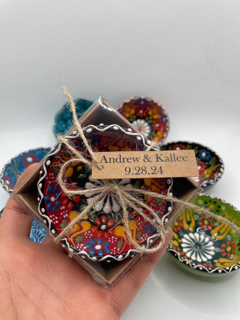 May include: A small, decorative ceramic bowl with a vibrant floral design in red, yellow, blue, and white. The bowl is packaged in a brown box and tied with twine. The tag reads "Andrew & Kallee 9.28.24".