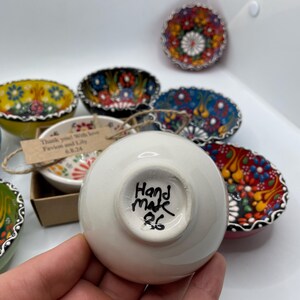 May include: A collection of small, hand-painted ceramic bowls with colorful floral designs. One bowl is held in a hand, showing the words "Hand made 26" on the bottom. Other bowls are visible in the background, each with unique patterns.