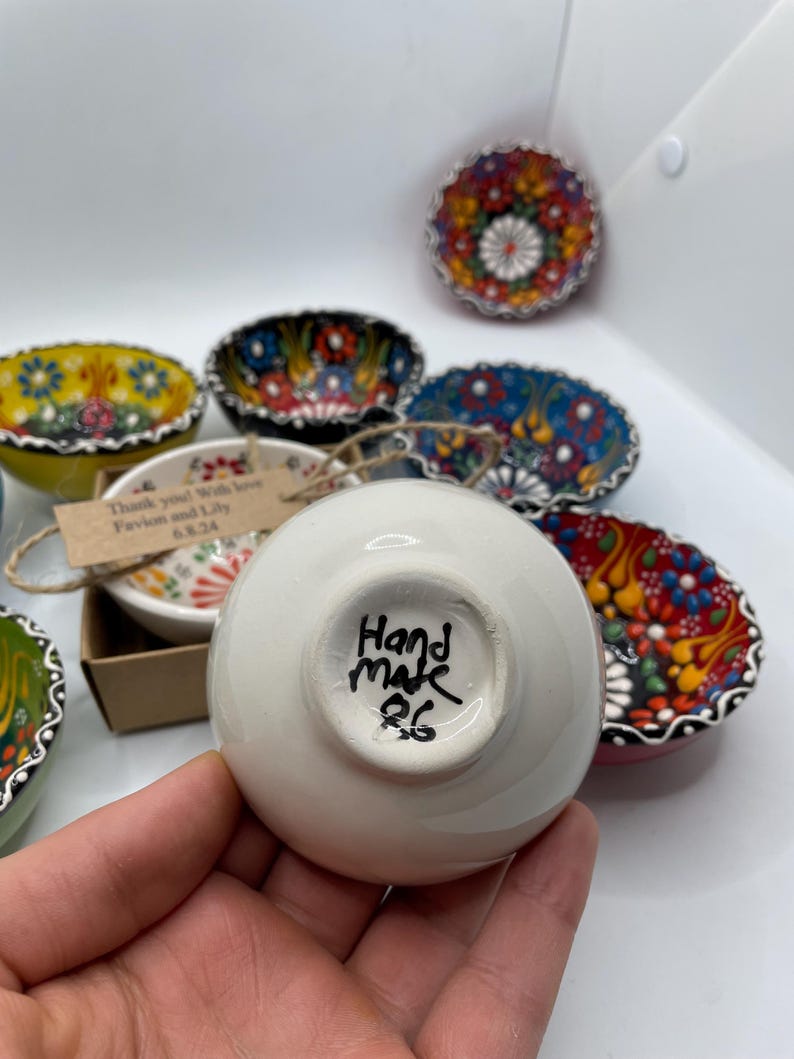 May include: A collection of small, hand-painted ceramic bowls with colorful floral designs. One bowl is held in a hand, showing the words "Hand made 26" on the bottom. Other bowls are visible in the background, each with unique patterns.