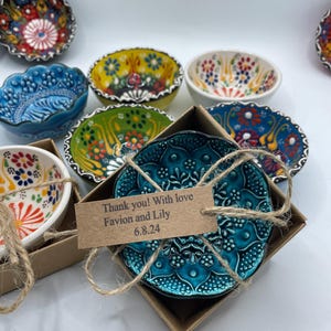 Op de afbeelding: Een verzameling kleine, decoratieve keramische kommen met ingewikkelde bloempatronen in verschillende kleuren. Een turquoise kom zit in een doos met een label met de tekst "Thank you! With love Favion and Lily 6.8.24". De kommen hebben geschulpte randen.