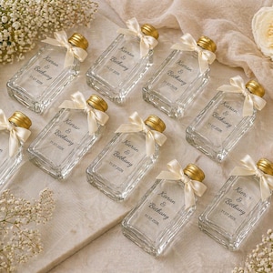 May include: A collection of clear glass bottles with gold-colored lids, each adorned with a cream-colored ribbon. The bottles are personalized with names and a date. The arrangement includes white flowers and a soft, neutral background.