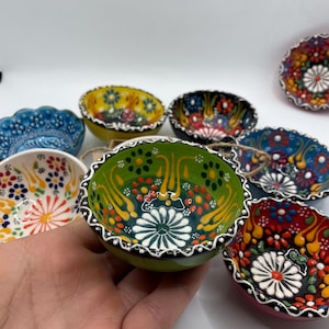 May include: A collection of small, hand-painted ceramic bowls with intricate floral designs. The bowls feature a variety of colors, including green, yellow, blue, and red, with white and black accents. The bowls are likely for serving dips or small portions.