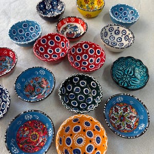 May include: A collection of small, decorative ceramic bowls in various colors, including blue, red, yellow, and orange. Each bowl features intricate hand-painted designs, such as floral patterns and eye-like motifs. The bowls have scalloped edges and are arranged on a white surface.