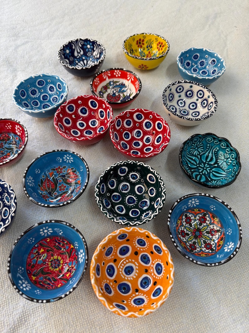 May include: A collection of small, decorative ceramic bowls in various colors, including blue, red, yellow, and orange. Each bowl features intricate hand-painted designs, such as floral patterns and eye-like motifs. The bowls have scalloped edges and are arranged on a white surface.