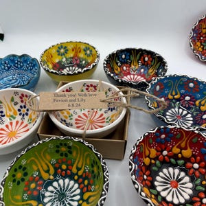 May include: A collection of small, hand-painted ceramic bowls with intricate floral designs in various colors, including blue, yellow, red, and green. The bowls have scalloped edges and are arranged on a white surface. One bowl has a tag that reads "Thank you! With love Favion and Lily 6.8.24."