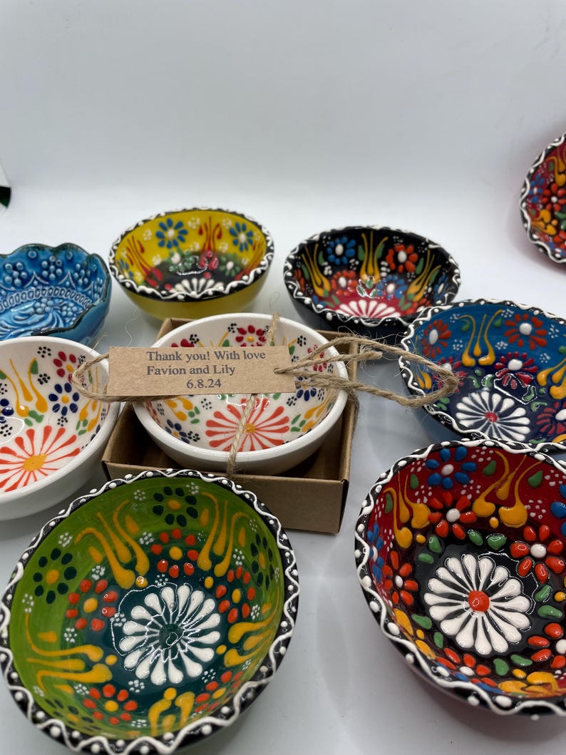 May include: A collection of small, hand-painted ceramic bowls with intricate floral designs in various colors, including blue, yellow, red, and green. The bowls have scalloped edges and are arranged on a white surface. One bowl has a tag that reads "Thank you! With love Favion and Lily 6.8.24."