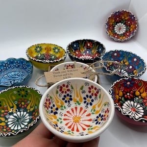 May include: A collection of small, hand-painted ceramic bowls with colorful floral designs. The bowls feature a variety of colors, including red, blue, yellow, and green, with intricate patterns. A small tag reads "Thank you! With love Favion and Lily 6.8.24."