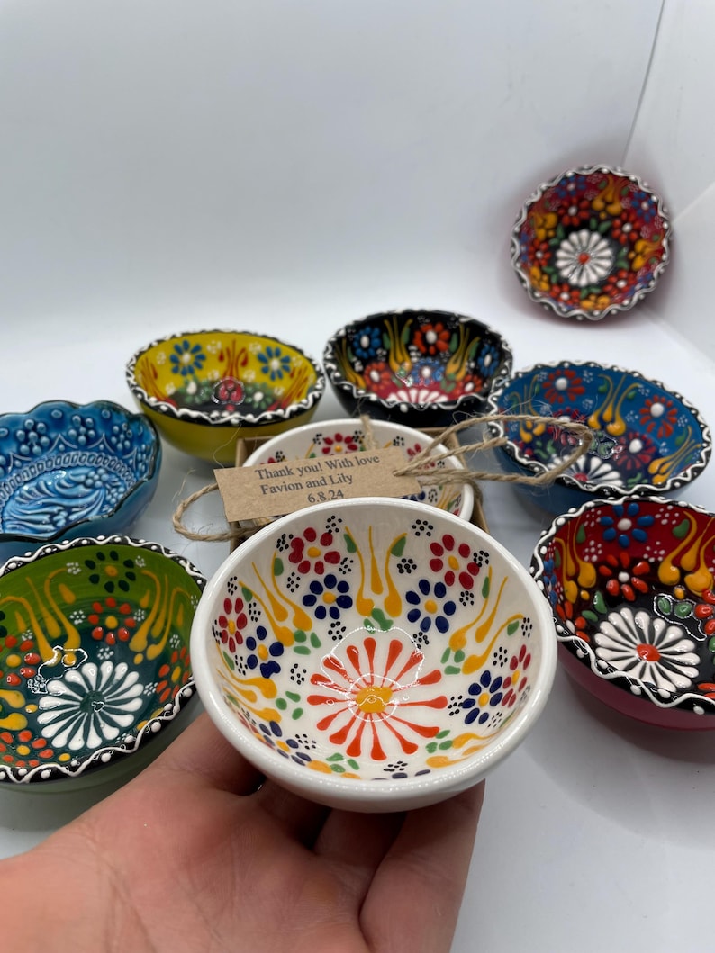 May include: A collection of small, hand-painted ceramic bowls with colorful floral designs. The bowls feature a variety of colors, including red, blue, yellow, and green, with intricate patterns. A small tag reads "Thank you! With love Favion and Lily 6.8.24."