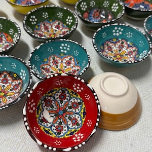 Peut inclure: Ensemble de petits bols en céramique peints à la main, ornés de motifs floraux colorés. Les bols présentent diverses couleurs, notamment le rouge, le vert, le jaune et le bleu, avec des bordures blanches. Ils sont probablement destinés à servir des sauces ou de petites portions.