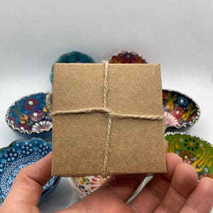 May include: A small, square, brown cardboard box tied with twine. The box is held in a hand, with several decorative, colorful ceramic bowls in the background. The bowls have floral and geometric patterns.