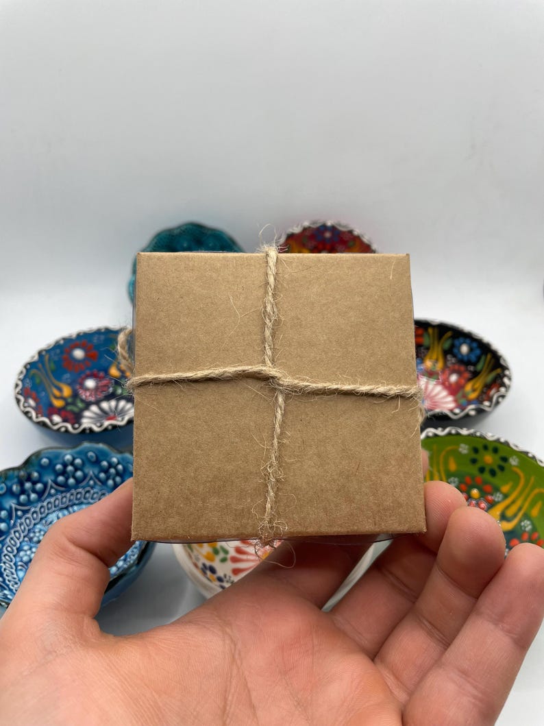 May include: A small, square, brown cardboard box tied with twine. The box is held in a hand, with several decorative, colorful ceramic bowls in the background. The bowls have floral and geometric patterns.