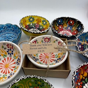 May include: A collection of colorful, hand-painted ceramic bowls with floral designs. One bowl is in a small cardboard box with a tag that reads "Thank you! With love Favion and Lily 6.8.24."
