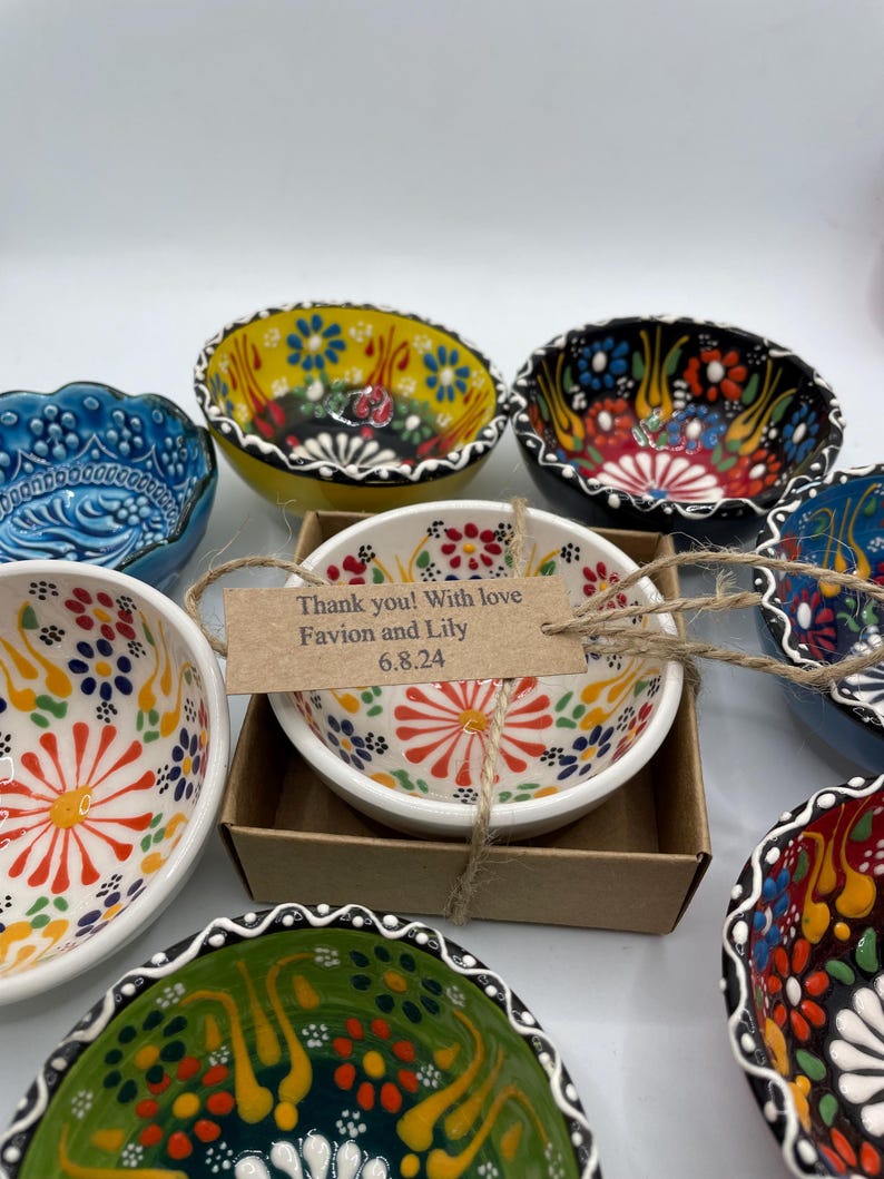 May include: A collection of colorful, hand-painted ceramic bowls with floral designs. One bowl is in a small cardboard box with a tag that reads "Thank you! With love Favion and Lily 6.8.24."