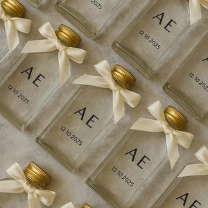 Pode incluir: Frascos de vidro transparente com tampas douradas e laços creme. Cada frasco está rotulado com "AE" e a data "12 10 2025" em preto. Dispostos sobre uma superfície clara.