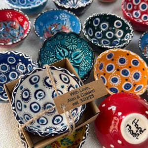May include: A selection of small ceramic bowls in shades of blue, red, green, and orange. The bowls display detailed patterns and designs, some with eye-shaped motifs. One bowl is packaged with a tag that reads "With Love, Alan & Colleen est. 3/22/25."