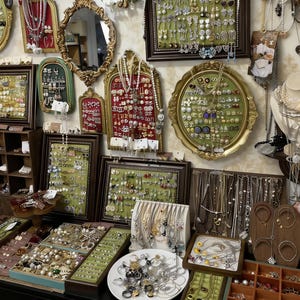May include: A shop display showcasing a variety of jewellery items. Necklaces, earrings, and bracelets are presented on trays, in framed displays, and hanging. The pieces incorporate pearls, gemstones, and diverse metal finishes. The overall style is vintage and ornate.