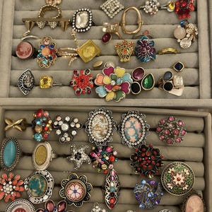 May include: A selection of rings in a grey jewellery tray. The rings have diverse designs, including floral, geometric, and gemstone embellishments. Colours range from vibrant reds and blues to neutral tones, showcasing a variety of styles and materials.